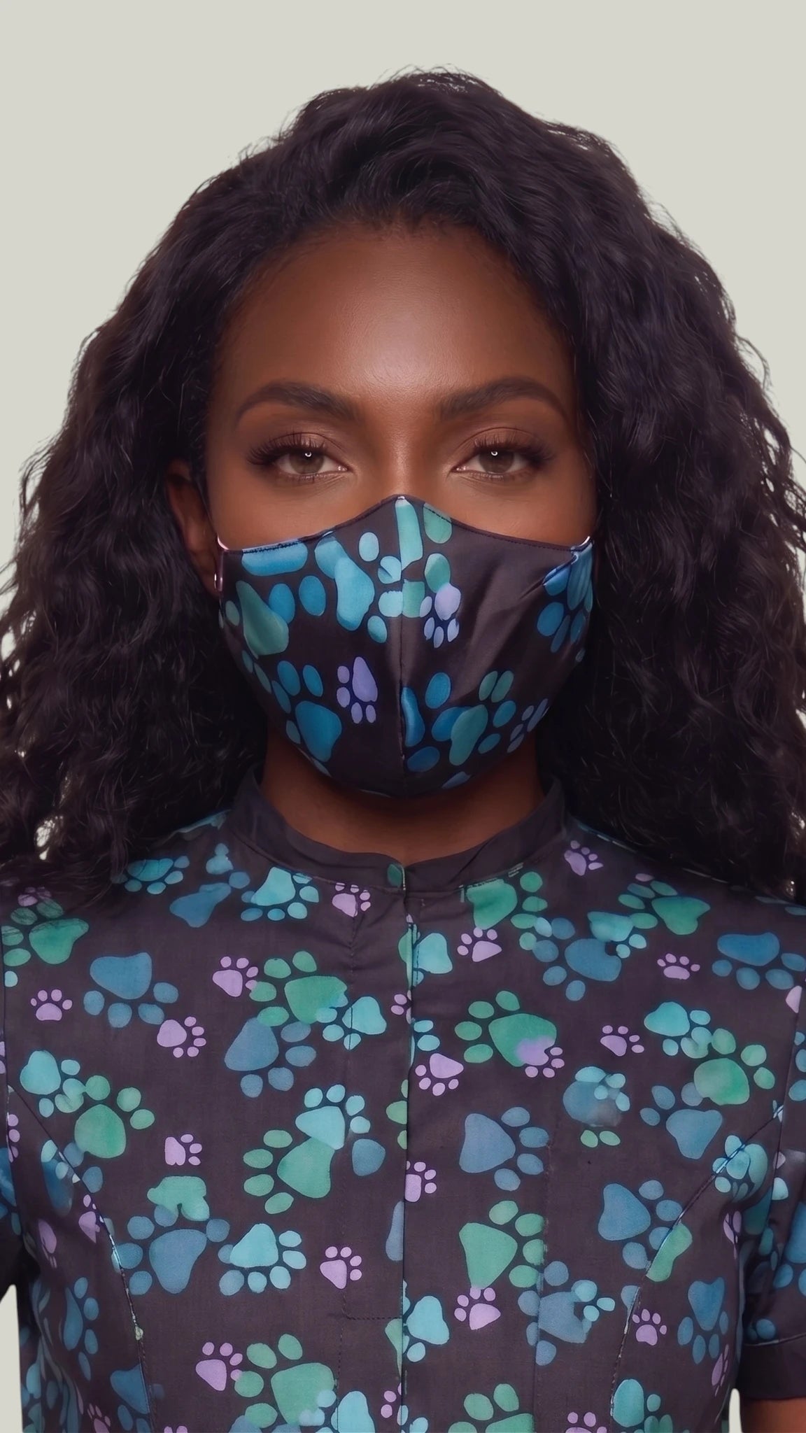 Close-up front view of a woman with long wavy dark hair modeling the Ladybird Line 24 pet grooming face mask in a blue paw print pattern on black fabric, paired with a matching short-sleeve grooming top against a light gray background.