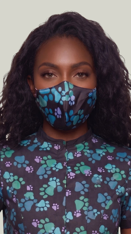 Close-up front view of a woman with long wavy dark hair modeling the Ladybird Line 24 pet grooming face mask in a blue paw print pattern on black fabric, paired with a matching short-sleeve grooming top against a light gray background.