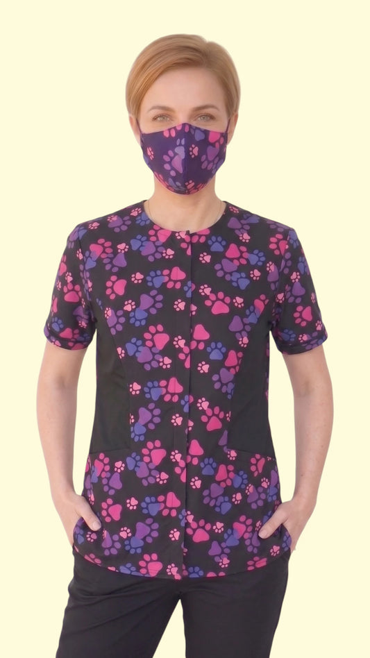 Front view of a woman with short blonde hair modeling the Ladybird Line 27 pink and purple paw print pet grooming face mask, paired with a matching short-sleeve grooming top against a pale yellow background.