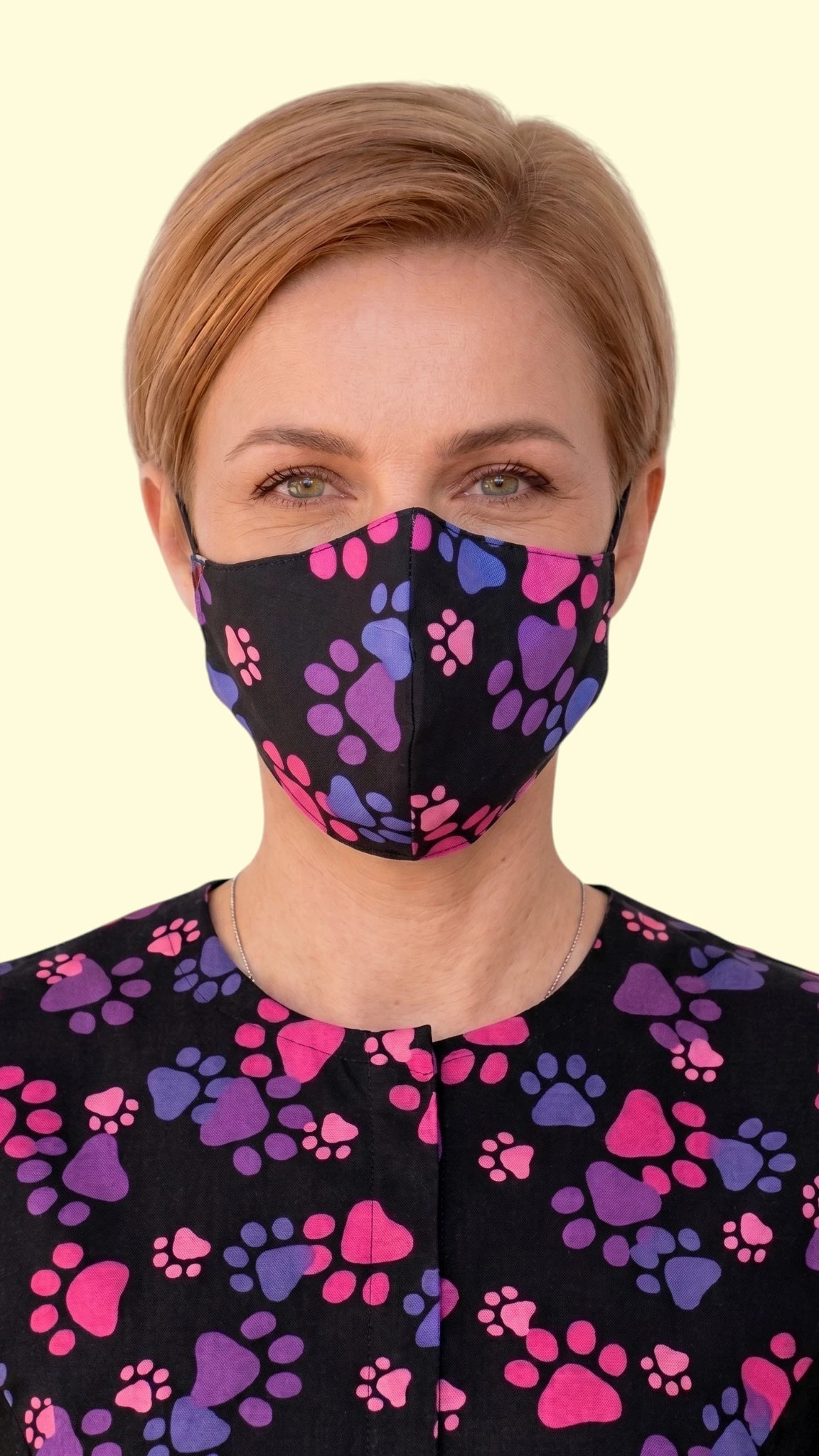 Close-up front view of a woman with short blonde hair modeling the Ladybird Line 27 pet grooming face mask in a vibrant pink and purple paw print pattern on black fabric, paired with a matching short-sleeve grooming top against a pale yellow background.