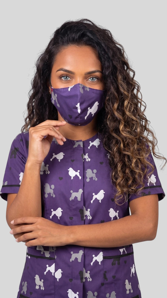 Front view of a woman with long curly brunette hair modeling the Ladybird Line 29 purple poodle pet grooming face mask, paired with a matching purple poodle grooming smock top against a gray studio background.