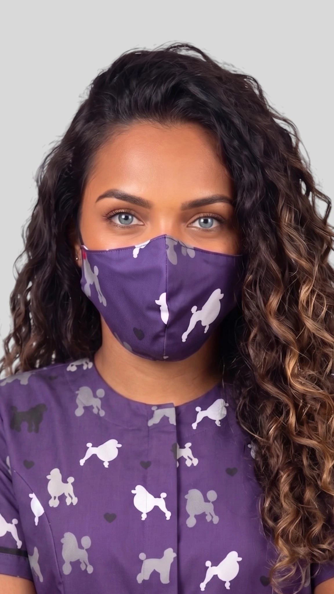 Close-up front view of a woman with long curly brown hair modeling the Ladybird Line 29 purple poodle pet grooming face mask, paired with a matching purple poodle grooming smock top against a gray studio background.