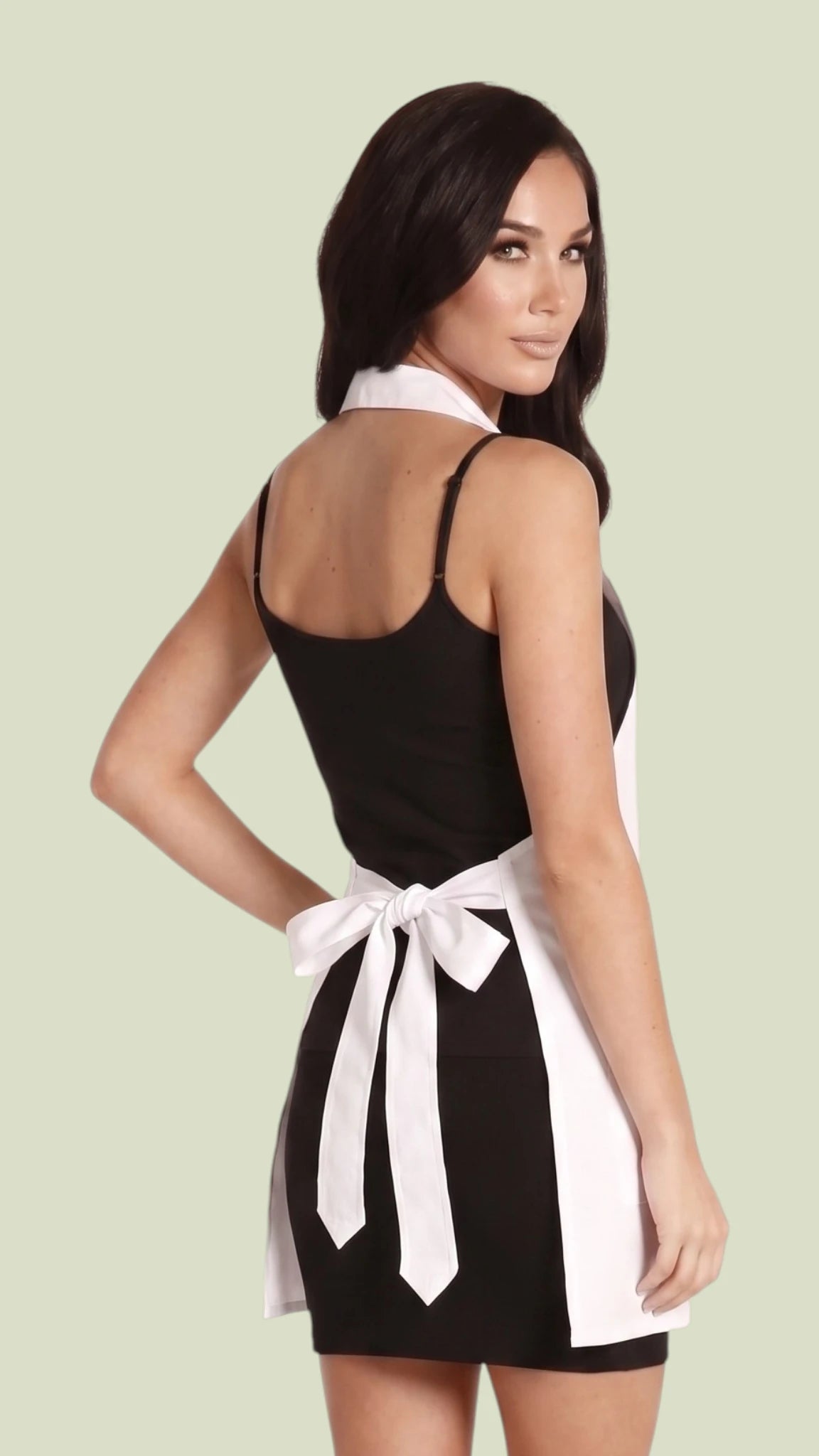 Back three-quarter view of a brunette woman modeling the Ladybird Line 290-W white pro jacket stylist apron, highlighting the tied white waist bow and halter-style collar over a black mini dress against a solid light beige background.