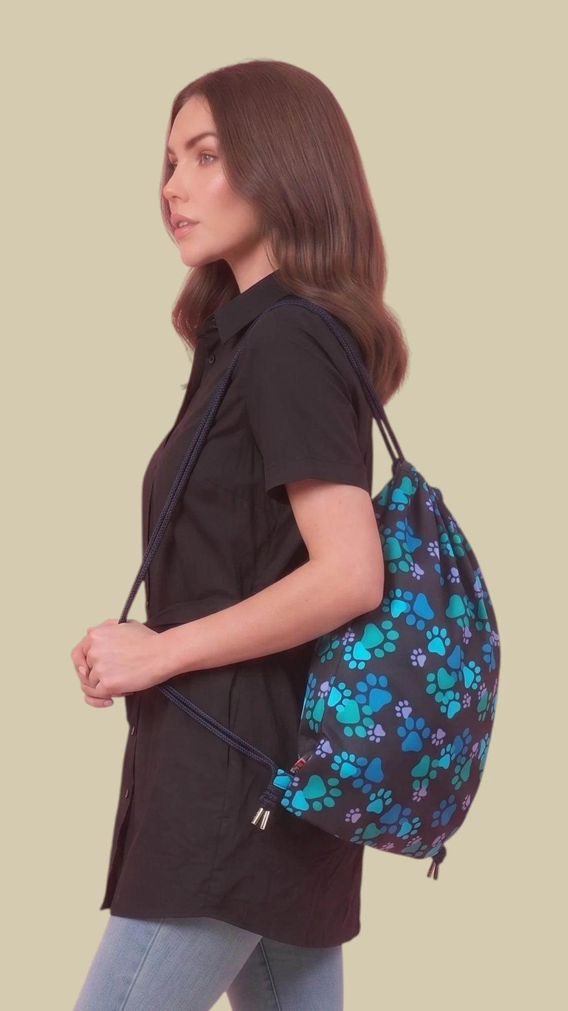 Side profile view of a woman with long brunette hair carrying a black Ladybird Line 320 drawstring backpack patterned with blue and teal paw prints over her shoulder, paired with a black short-sleeve tunic and blue jeans against a solid beige background.