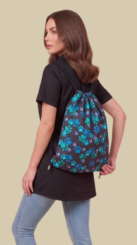 Back three-quarter view of a woman with long brunette hair modeling the Ladybird Line 320 drawstring backpack featuring an all-over blue and teal paw print pattern, paired with a black short-sleeve tunic top and light blue jeans against a solid light beige background.