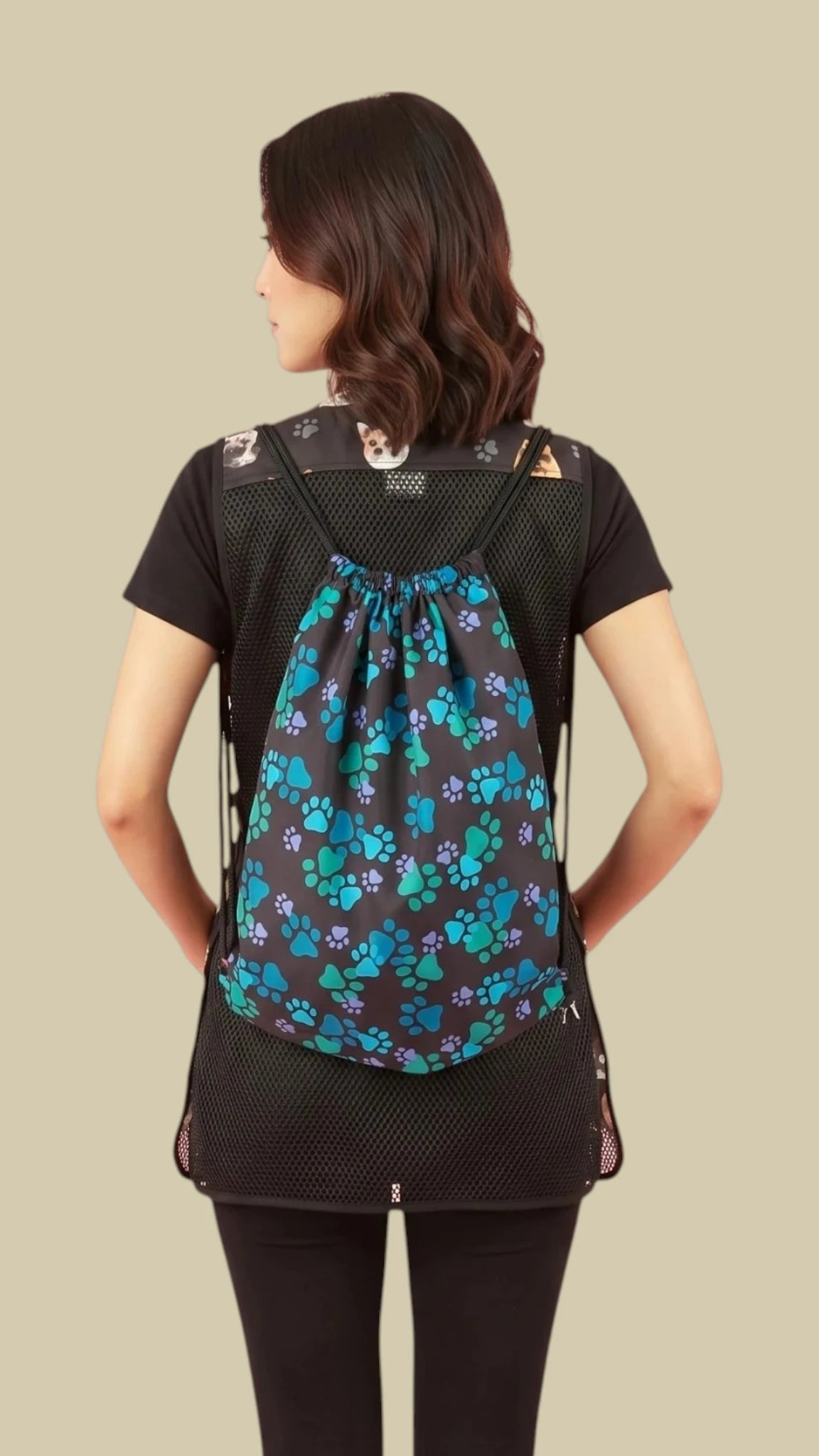 Full back view of a woman with dark hair wearing a black short-sleeve shirt and black leggings, wearing the Ladybird Line 320 cinch drawstring backpack featuring an all-over blue and teal paw print pattern against a solid light beige background.