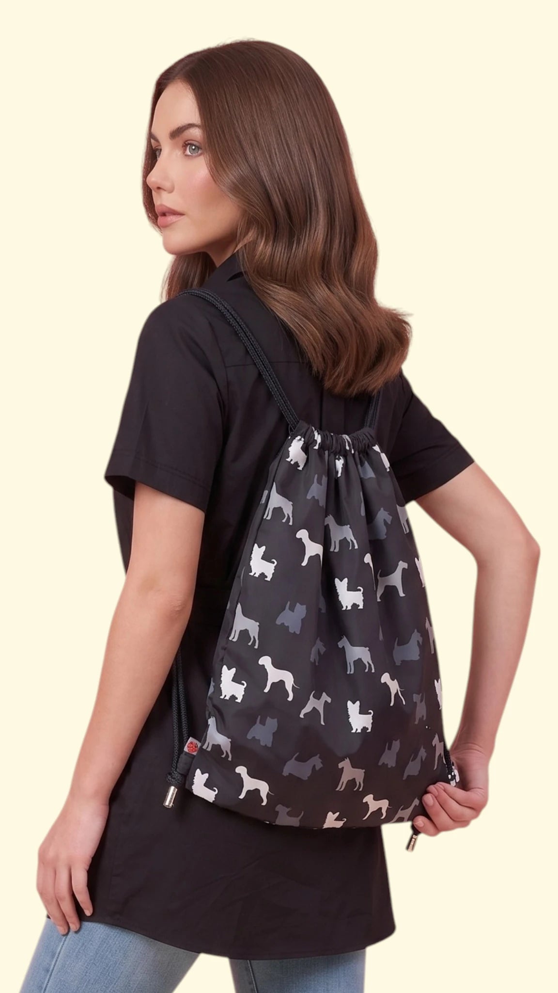 Side and back view of a woman modeling the Ladybird Line 340 drawstring backpack featuring a black, grey, and white Scottish Terrier dog print, paired with a black short-sleeve tunic shirt and blue jeans against a solid off-white background.