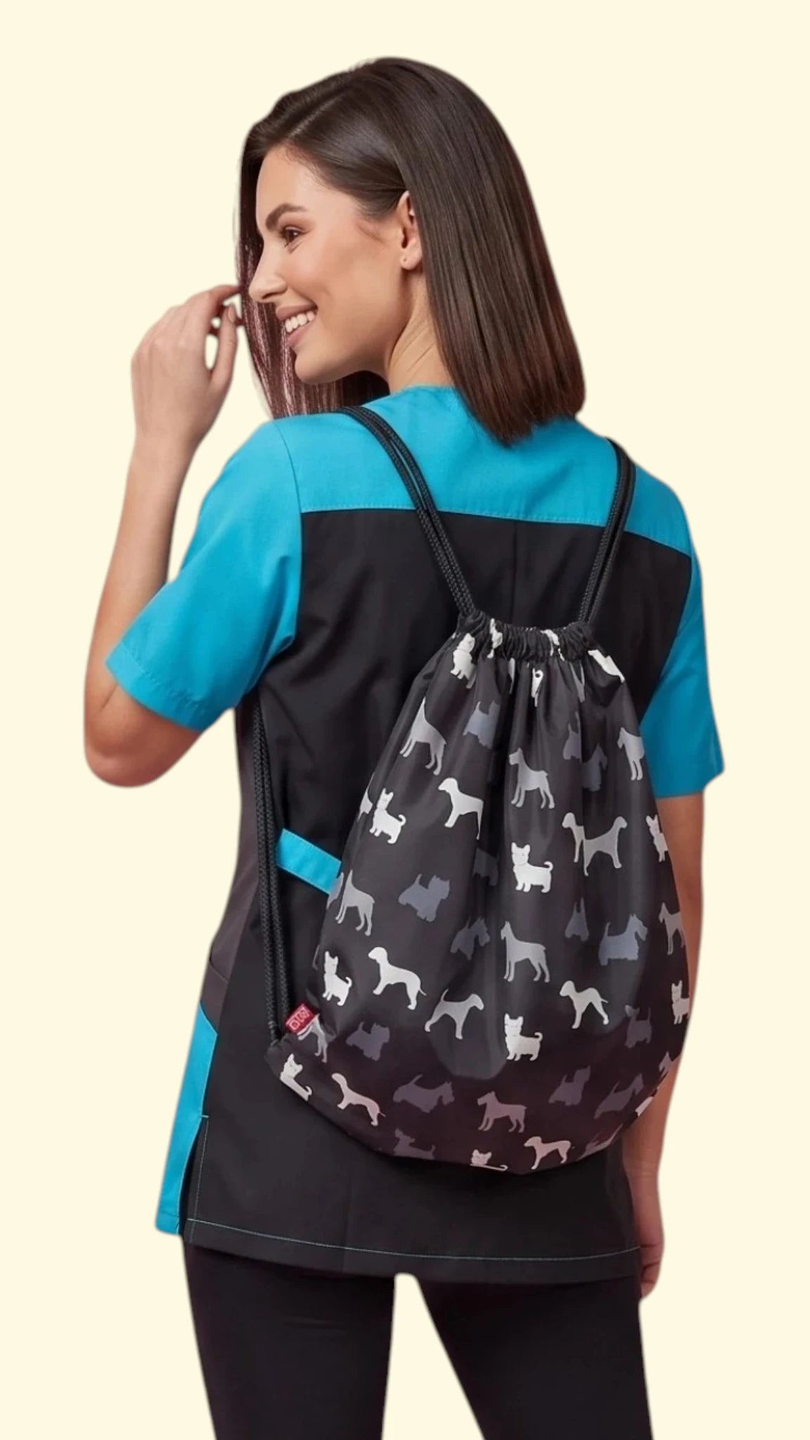 Back three-quarter view of a woman with dark hair wearing a black and teal grooming scrub tunic, carrying a black drawstring backpack patterned with Scottish Terrier dog silhouettes in white, grey, and black against a solid light cream background.