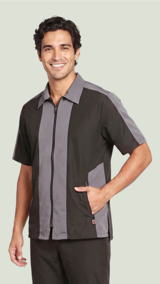 Front view of a smiling man modeling the Ladybird Line 418 men's short-sleeve barber and grooming jacket in black with charcoal gray vertical accent panels, structured collar, and hand in the side pocket against a solid light gray background.