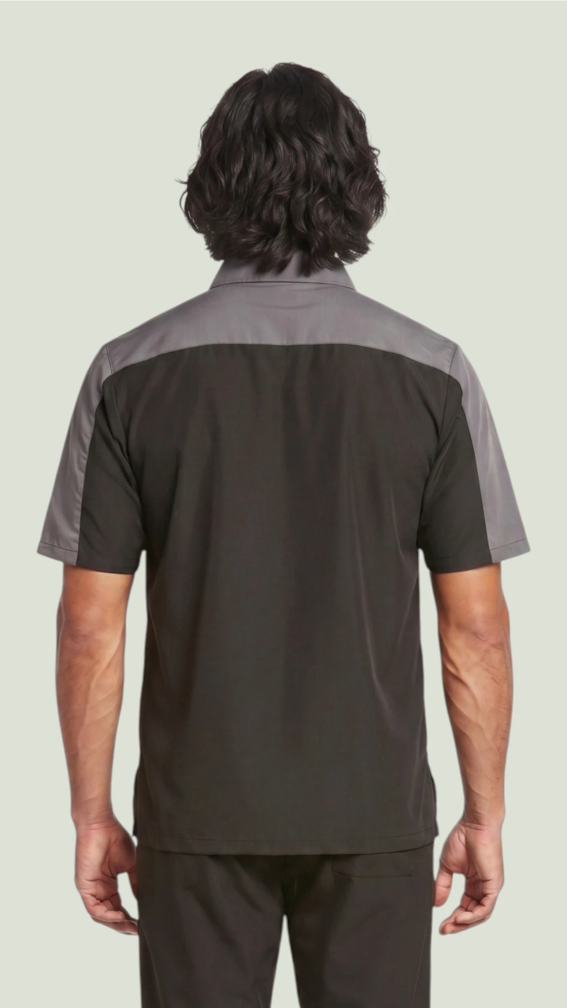 Back view of a dark-haired man modeling the Ladybird Line 418 men's short-sleeve barber and grooming jacket, featuring a black body with charcoal gray upper back shoulder panels, paired with dark trousers against a solid light gray background.