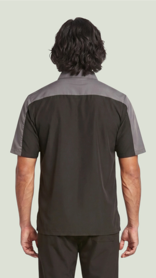Back view of a dark-haired man modeling the Ladybird Line 418 men's short-sleeve barber and grooming jacket, featuring a black body with charcoal gray upper back shoulder panels, paired with dark trousers against a solid light gray background.