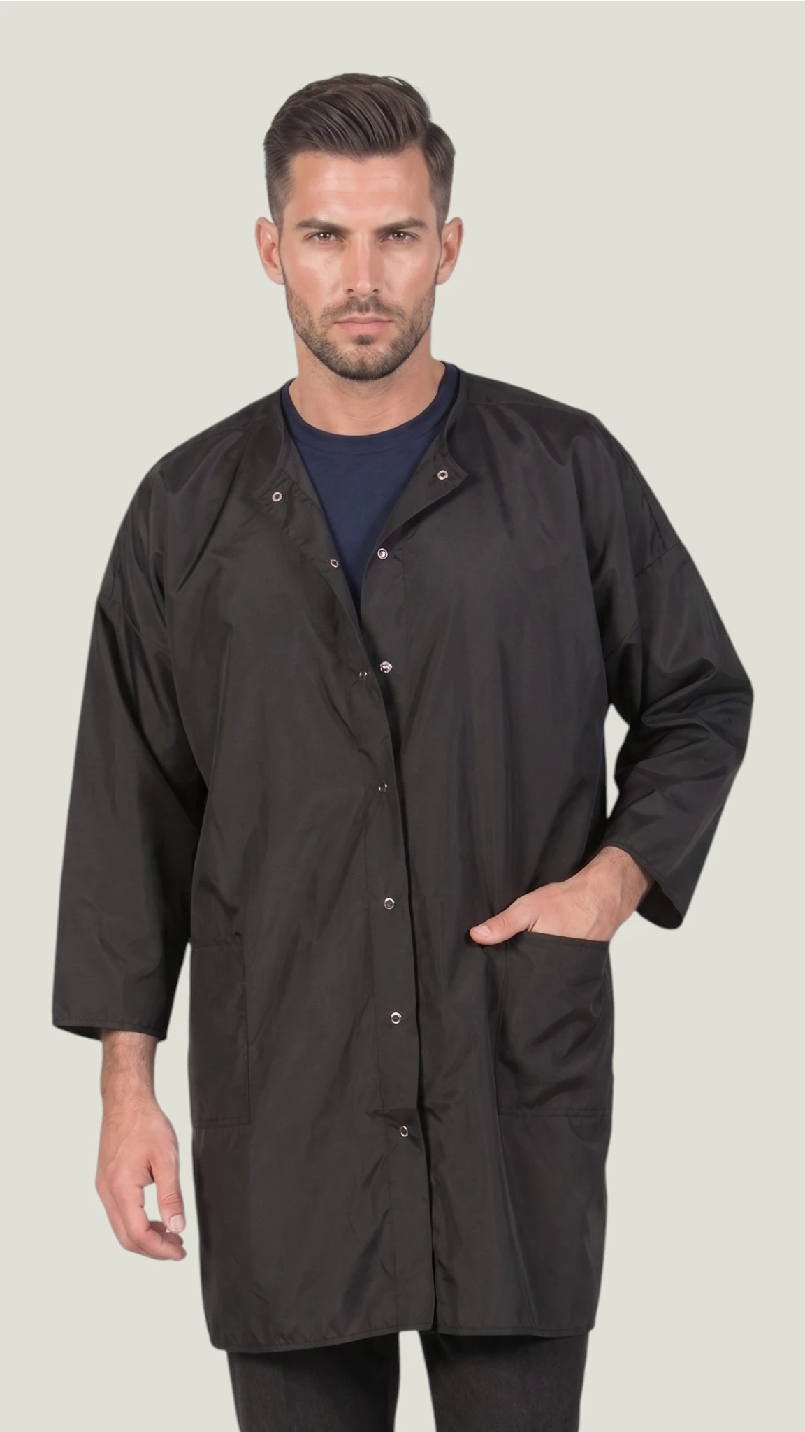 Front view of a man modeling the Ladybird Line 428 unisex black water and bleach-resistant groomer and client big shirt smock gown with snap button closure and 3/4 sleeves against a solid light gray background.