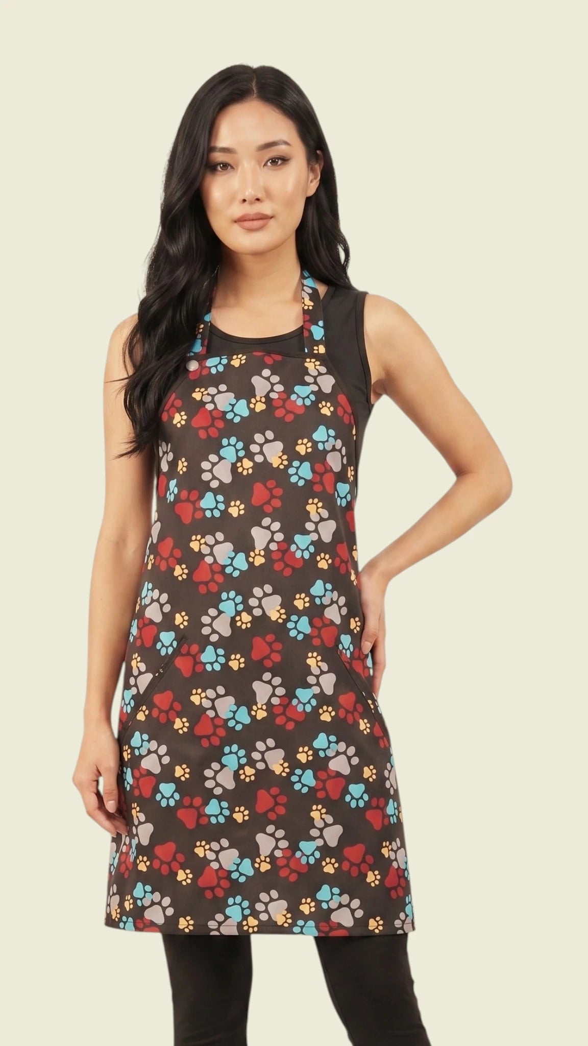 Three-quarter length front view of a woman with long dark hair posing with one hand on her hip, modeling the Ladybird Line 601 waterproof pet grooming apron in a multicolor paw print pattern over a black tank top and leggings against a light cream background.