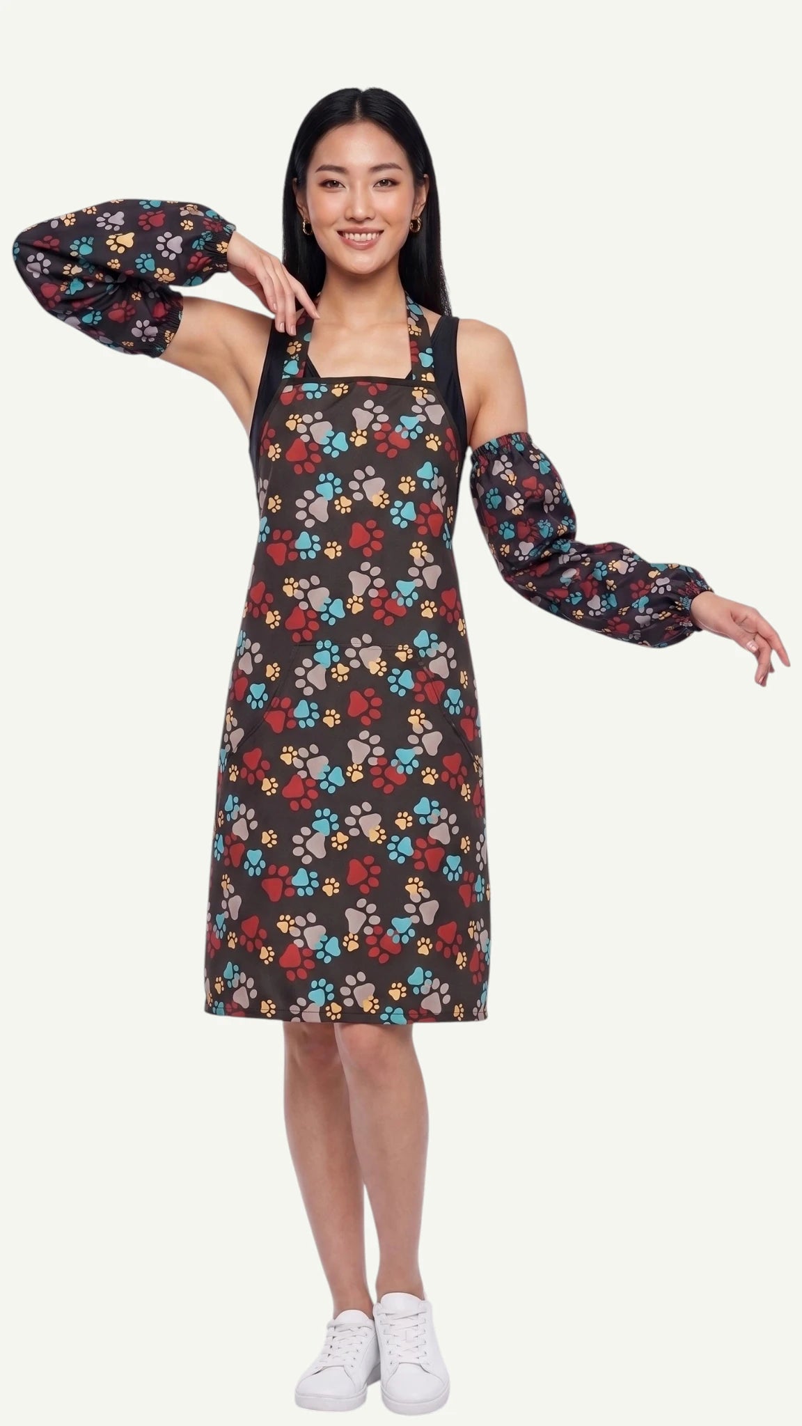 Full-length front view of a smiling woman modeling the Ladybird Line 603 waterproof paw print dog bathing sleeves, paired with a matching paw print pet grooming apron dress against an off-white background.