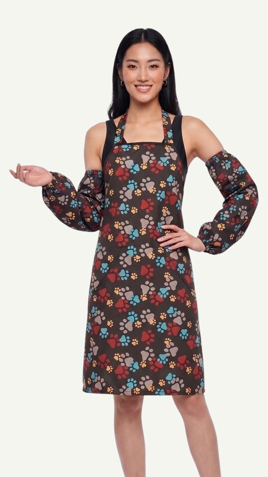 Front view of a smiling dark-haired woman modeling the Ladybird Line 603 waterproof paw print dog bathing sleeves, paired with a matching paw print pet grooming apron against a plain light background.