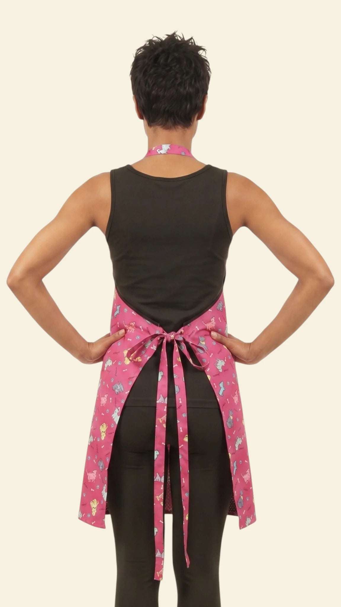 Rear view of a woman with short dark hair posing with hands on hips, modeling the Ladybird Line 609 waterproof pet grooming apron in vibrant pink with dog print, showing the tie-waist bow over a black sleeveless top and black leggings against a light beige background.