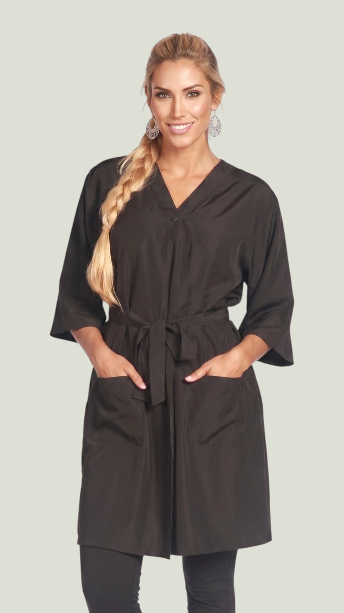 Front view of a smiling blonde woman with a side braid modeling the Ladybird Line 6306 black wrap-style client kimono robe with 3/4 sleeves, tied waist belt, and deep front pockets against an off-white background.