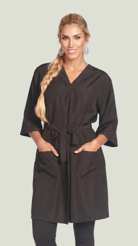 Front view of a smiling blonde woman with a side braid modeling the Ladybird Line 6306 black wrap-style client kimono robe with 3/4 sleeves, tied waist belt, and deep front pockets against an off-white background.