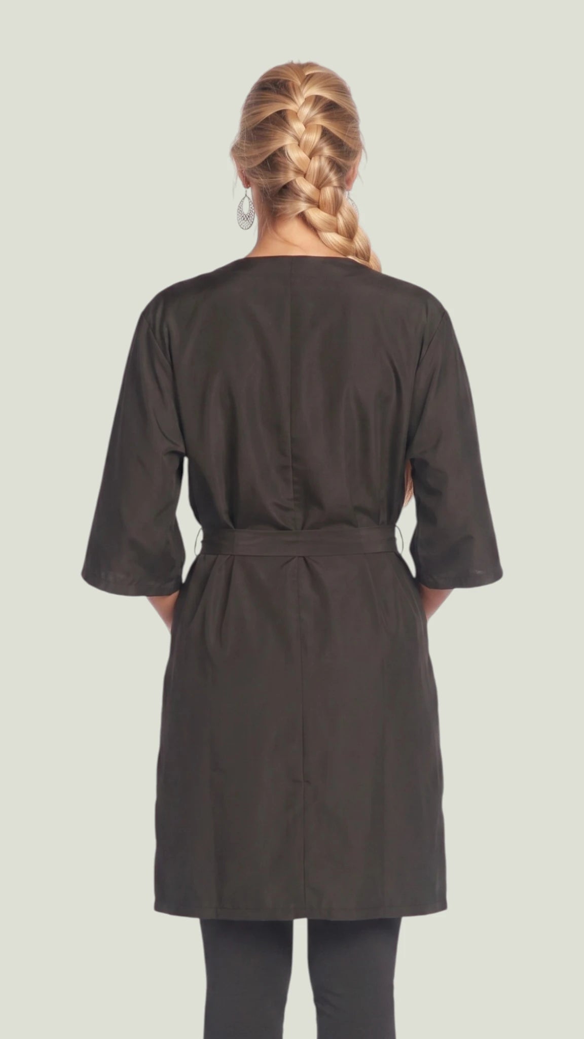 Back view of a woman with braided blonde hair modeling the Ladybird Line 6306 black client kimono robe, showing the tied waist belt, 3/4 sleeves, and knee-length hem, paired with dark leggings against a light studio background.