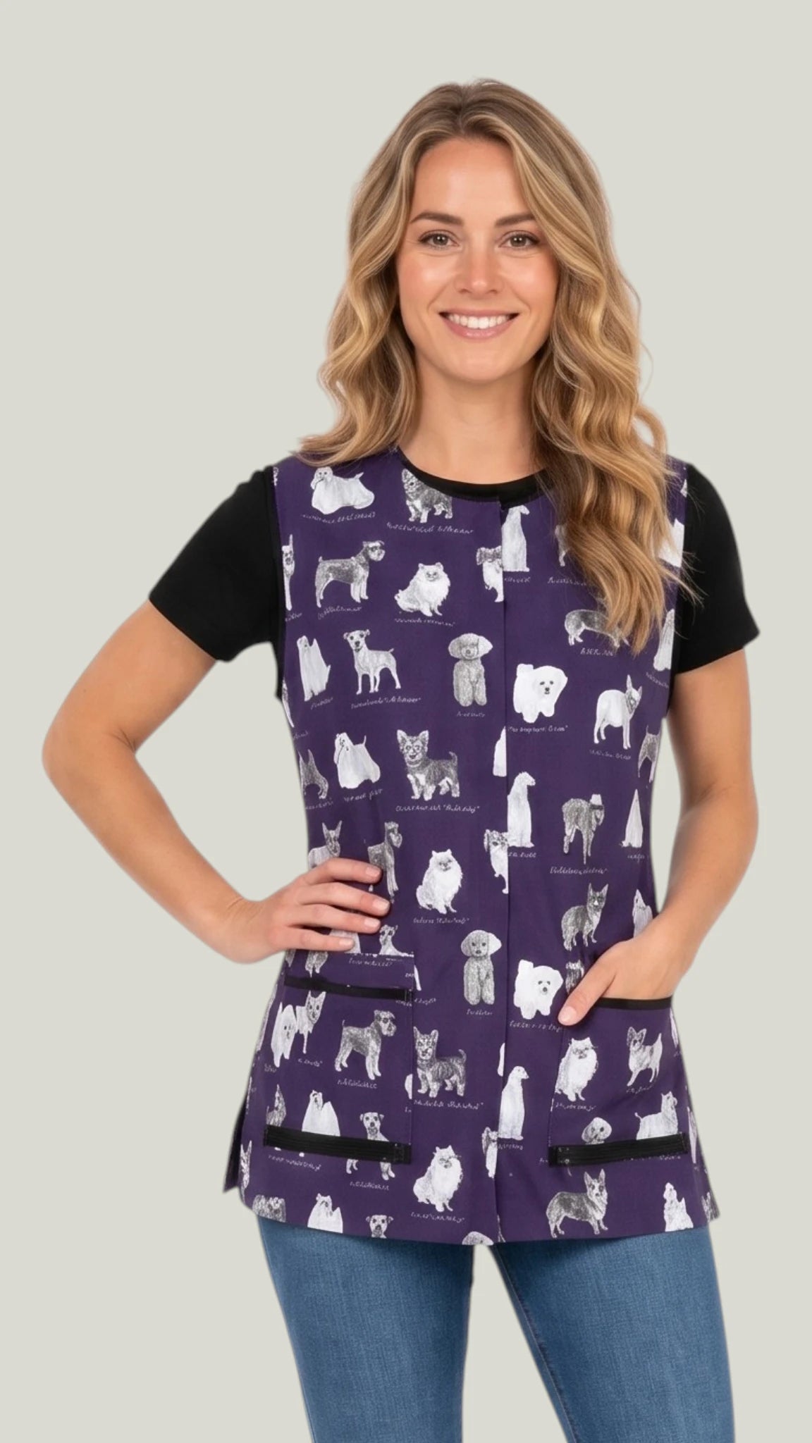 Three-quarter length front view of a blonde woman smiling with hand on hip modeling the Ladybird Line 740 Puppy Love sketch waterproof pet grooming vest in deep purple with puppy breed print, worn over a black t-shirt and jeans against a light gray background.