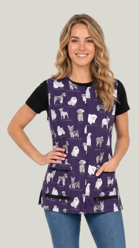 Three-quarter length front view of a blonde woman smiling with hand on hip modeling the Ladybird Line 740 Puppy Love sketch waterproof pet grooming vest in deep purple with puppy breed print, worn over a black t-shirt and jeans against a light gray background.