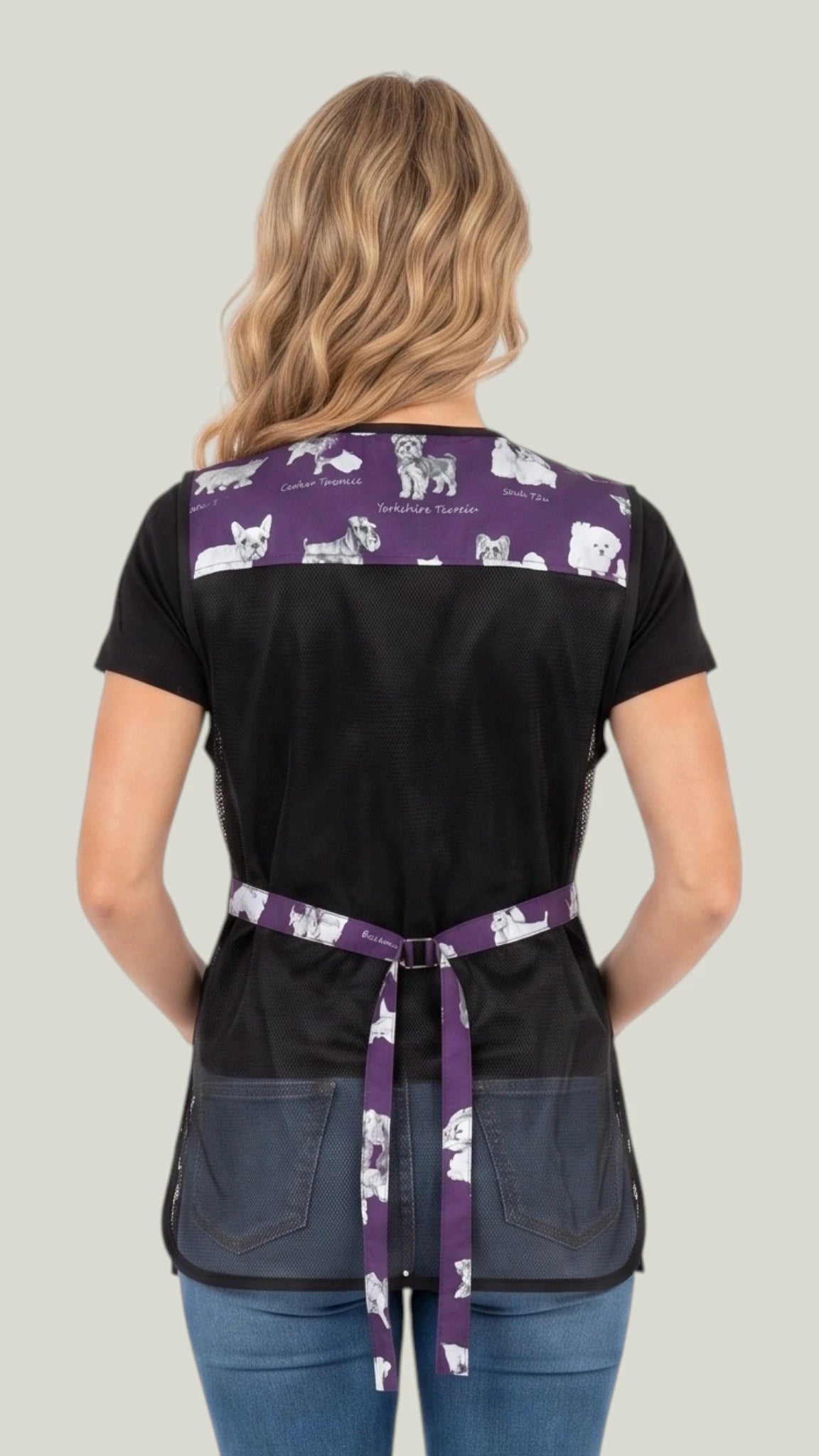 Rear view of a blonde woman modeling the Ladybird Line 740 Puppy Love sketch waterproof pet grooming vest, highlighting the breathable black mesh back, purple puppy print upper panel, and adjustable tie waist belts over blue jeans against a light gray background.