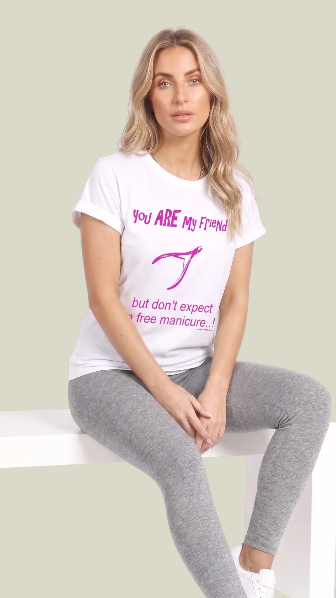 Full-body view of a blonde woman sitting on a white bench modeling a white short-sleeve cotton graphic t-shirt printed with pink text reading "You ARE My Friend but don't expect a free manicure...!" with a nail polish graphic, paired with heather grey leggings and white sneakers against a solid light sage green background.