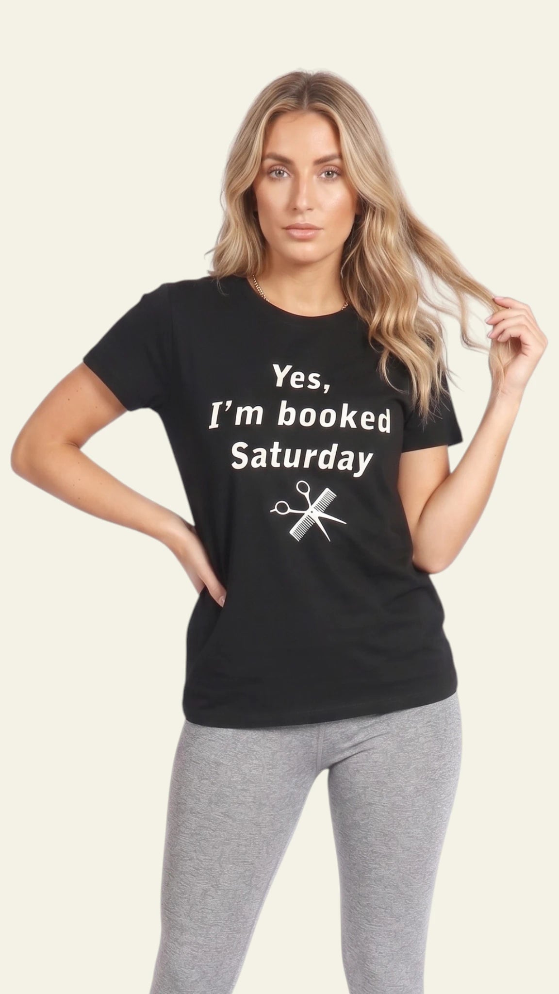 Front view of a blonde woman modeling a black short-sleeve t-shirt printed with white text reading "Yes, I'm booked Saturday" and a pair of scissors graphic, paired with light grey pants against a solid off-white background.
