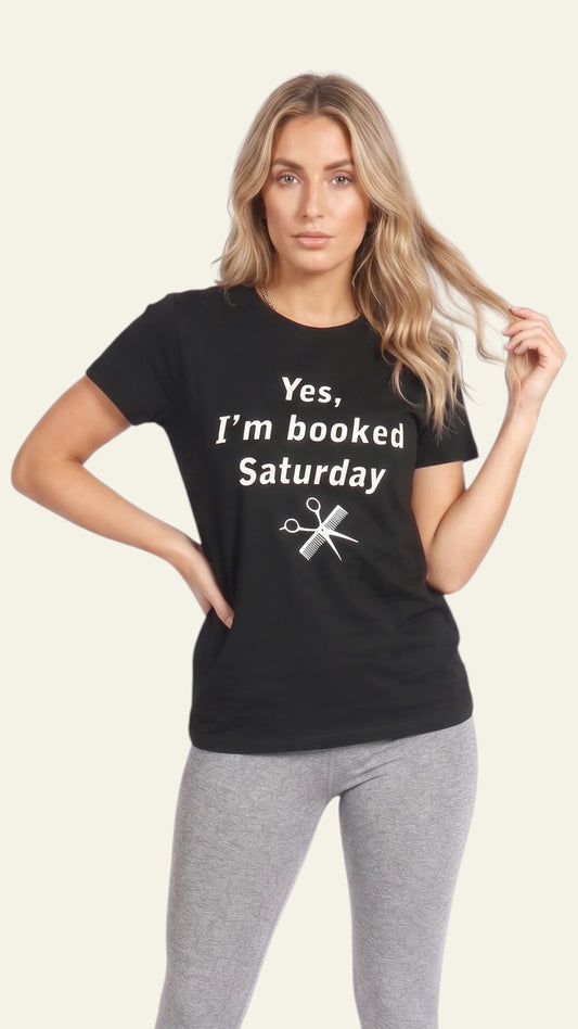 Front view of a blonde woman modeling a black short-sleeve t-shirt printed with white text reading "Yes, I'm booked Saturday" and a pair of scissors graphic, paired with light grey pants against a solid off-white background.