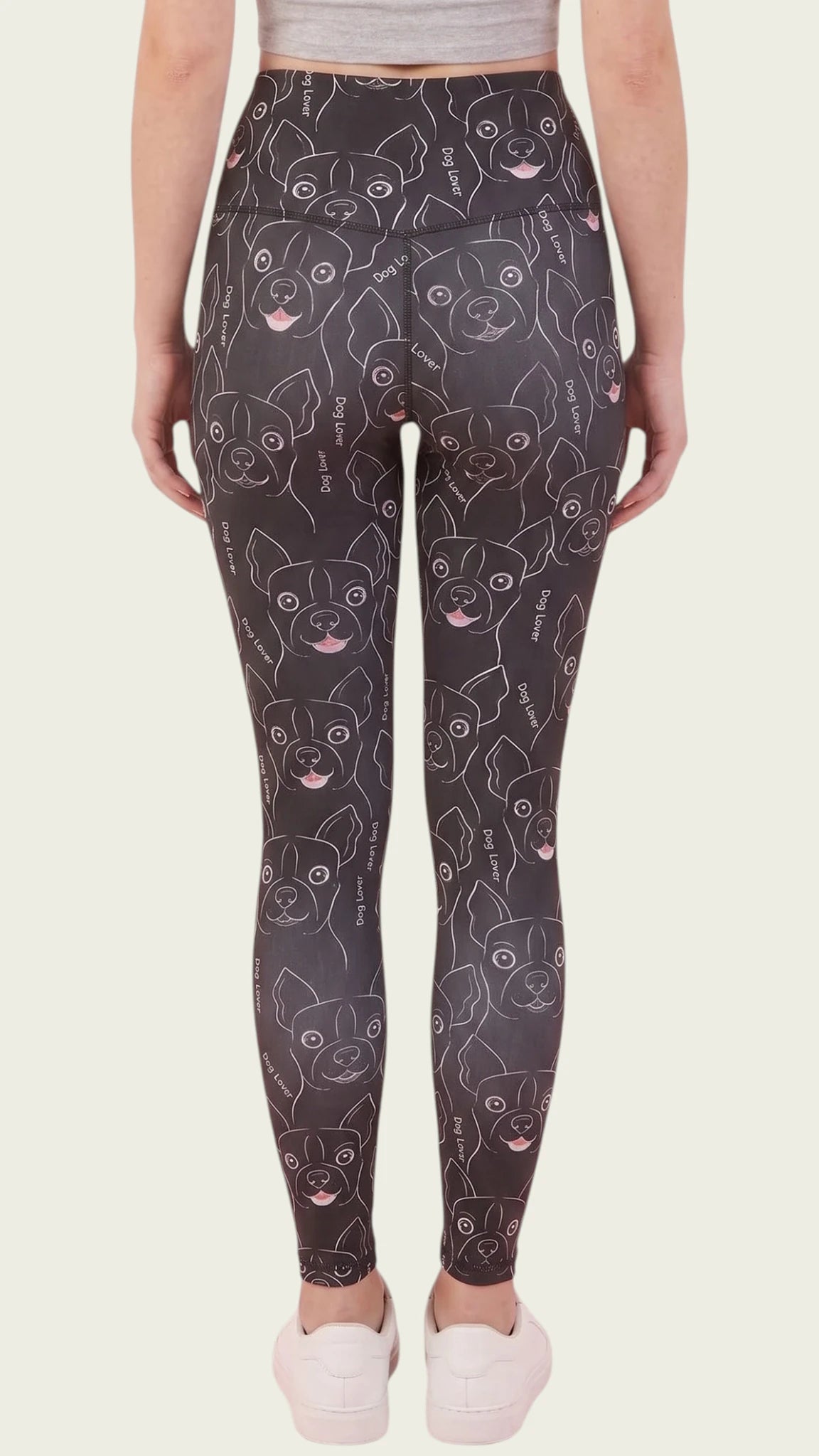 Back view of a woman modeling the Ladybird Line 124 Dog Lover Shadow Hair & Bleach Resistant Grooming Leggings featuring a black dog face line-art print, worn with white sneakers against a beige background.