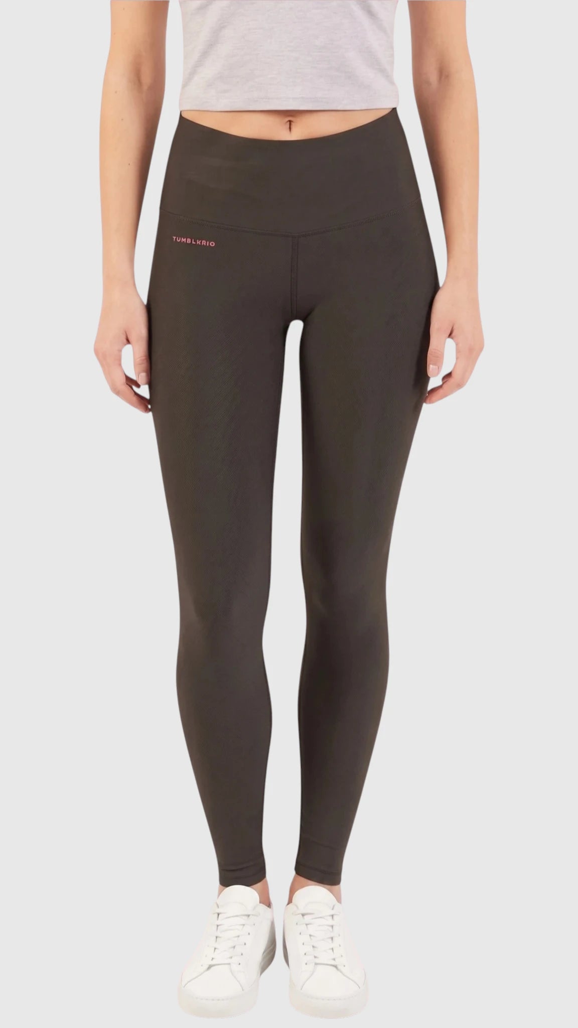 Front full-body view of a model wearing high-waisted black Ladybird Line 125 Black Shadow Grooming Leggings with a small silver logo on the left hip, paired with a light grey short-sleeve crop top and white sneakers, set against a light grey background.