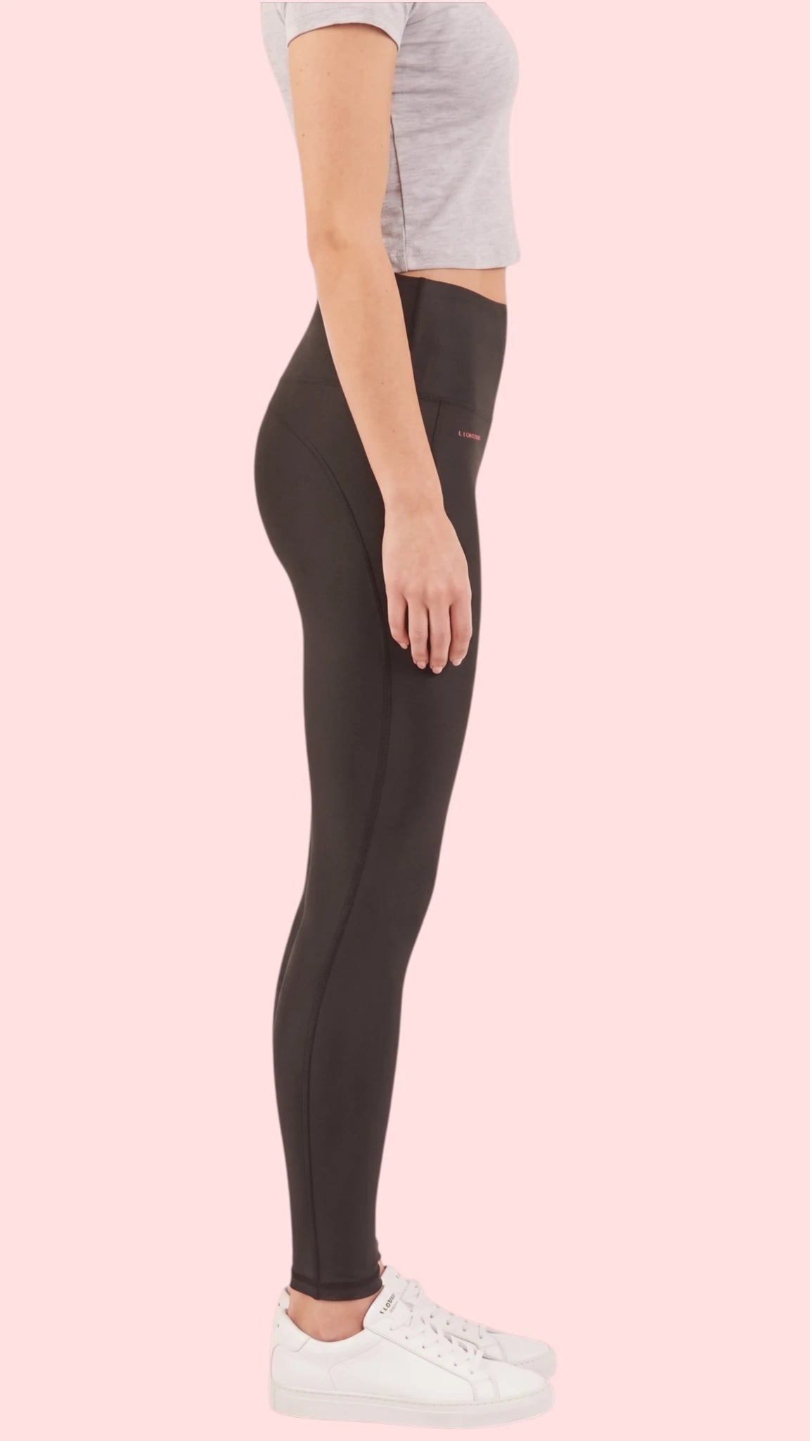 Side profile view of a woman modeling the Ladybird Line 125 Black Shadow Hair & Bleach Resistant Grooming Leggings, paired with a gray cropped t-shirt and white sneakers against a pale pink background.