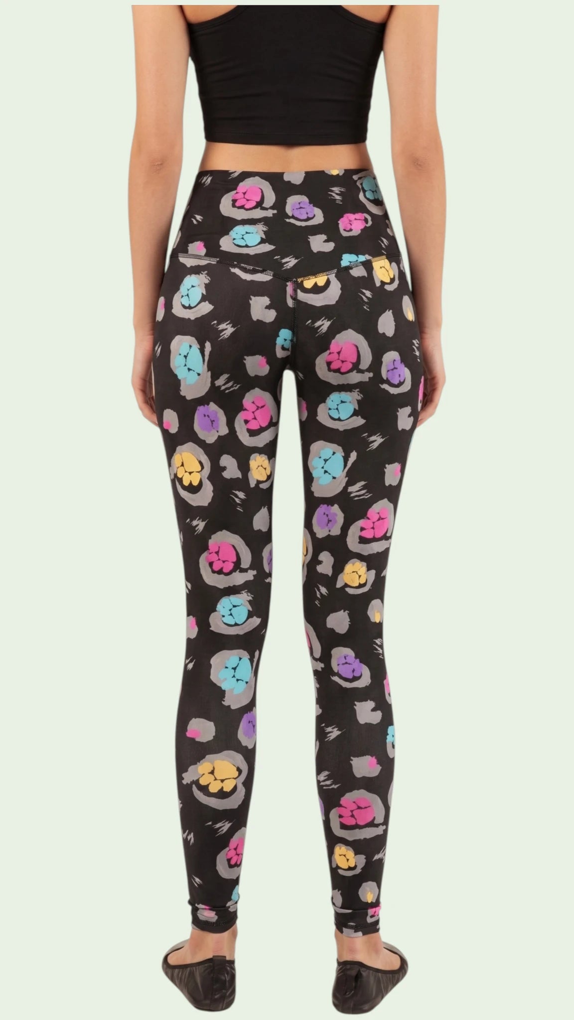 Back view of a woman modeling the Ladybird Line 126 Paw Print Shadow Hair & Bleach Resistant Grooming Leggings featuring a colorful pink, teal, purple, and yellow paw print pattern, styled with a black crop top and black flats against a light green background.