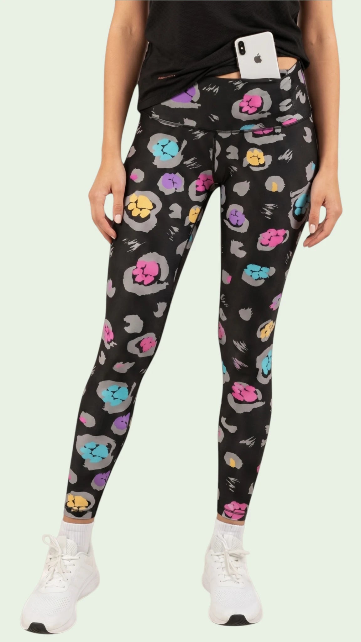 Front view of a woman modeling the Ladybird Line 126 Paw Print Shadow Hair & Bleach Resistant Grooming Leggings featuring a colorful pink, teal, purple, and yellow paw print pattern, styled with a black top and white sneakers against a pale background.