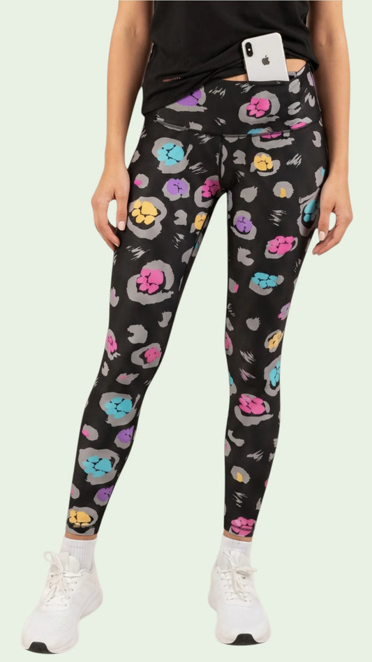 Front view of a woman modeling the Ladybird Line 126 Paw Print Shadow Hair & Bleach Resistant Grooming Leggings featuring a colorful pink, teal, purple, and yellow paw print pattern, styled with a black top and white sneakers against a pale background.