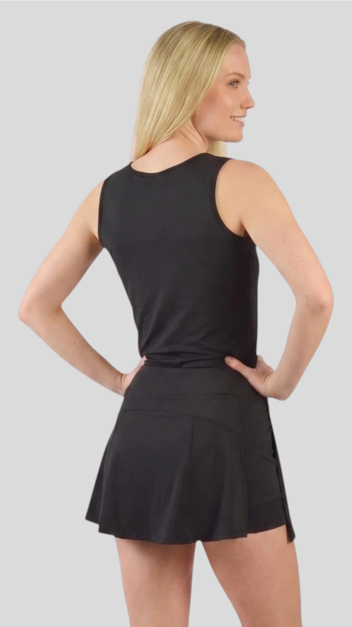 Back view of a blonde woman modeling the Ladybird Line 129 Black Shadow Hair & Bleach Resistant Grooming Skort with hands on her hips, paired with a matching black sleeveless tank top against a light gray background.