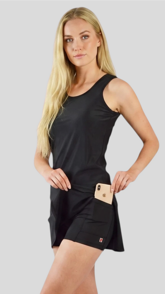 Front view of a woman modeling the Ladybird Line 129 Black Shadow Hair & Bleach Resistant Grooming Skort with a matching black tank top, featuring a functional side pocket holding a smartphone against a light gray background.