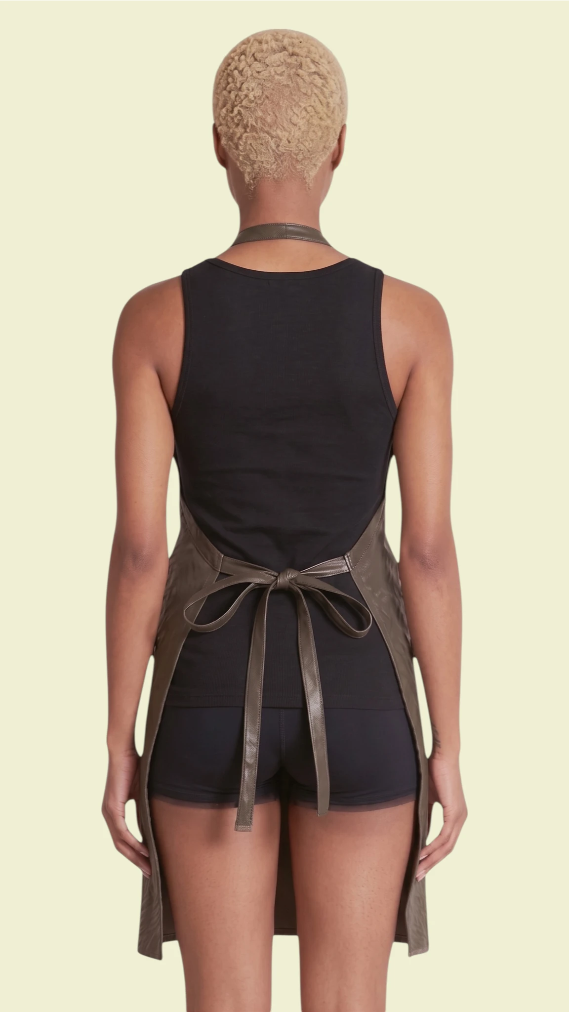 Back view of a woman with short blonde hair modeling the Ladybird Line 130 Black Leopard Print Hair & Bleach Resistant Grooming Apron, tied neatly at the waist over a black tank top and shorts against a pale yellow background.