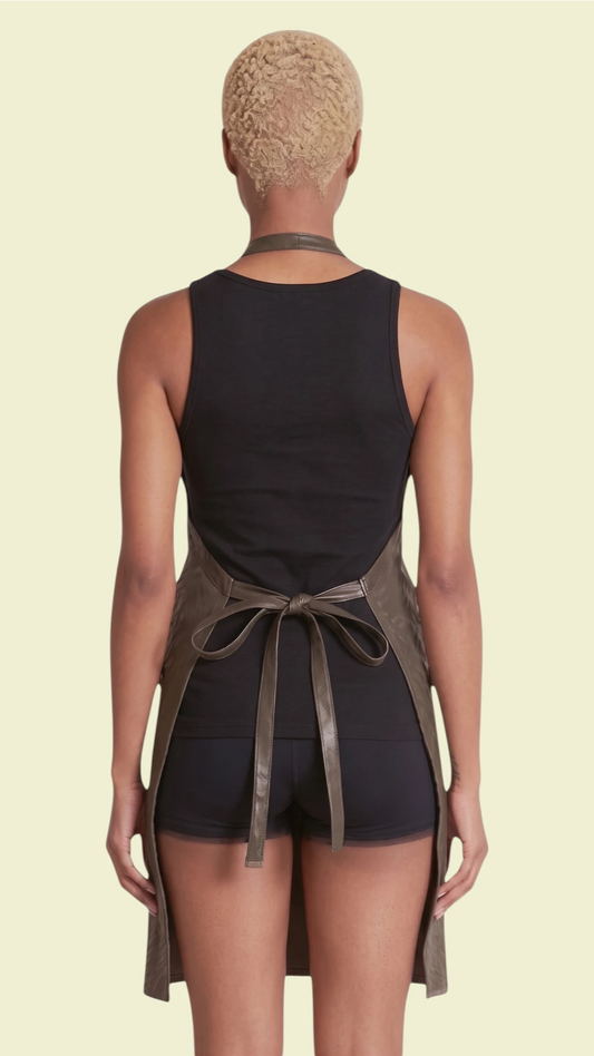 Back view of a woman with short blonde hair modeling the Ladybird Line 130 Black Leopard Print Hair & Bleach Resistant Grooming Apron, tied neatly at the waist over a black tank top and shorts against a pale yellow background.