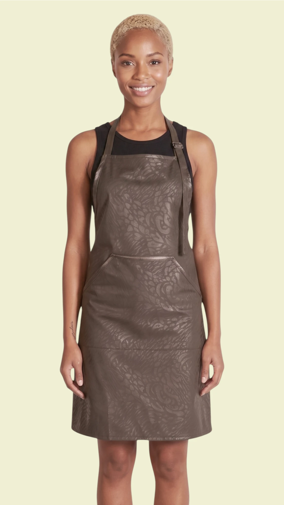 Front view of a woman modeling the Ladybird Line 130 Black Leopard Print Hair & Bleach Resistant Grooming Apron, featuring a subtle black leopard pattern, deep front pocket, and adjustable neck strap, worn over a black top against a pale yellow background.