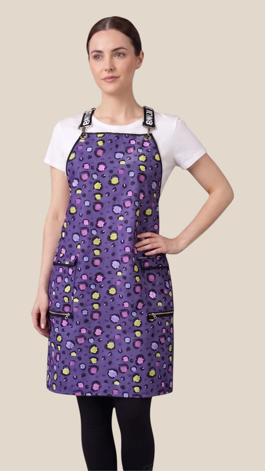 Front view of a woman modeling the Ladybird Line 139 Lilac Mini Paw Print Waterproof Signature Strap Crossover Apron, featuring a purple mini paw print pattern, deep zippered pockets, and branded crossover straps, worn over a white t-shirt and black pants against a light beige background.
