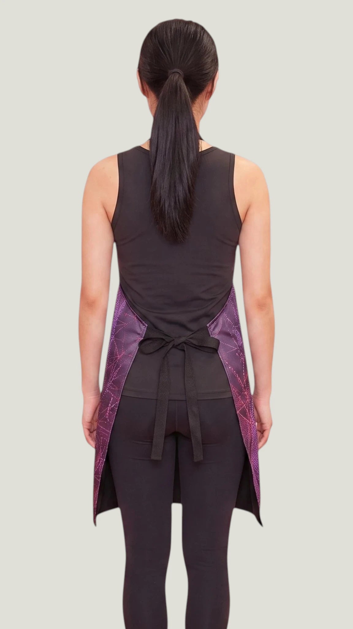 Back view of an Asian woman with a dark ponytail modeling the Ladybird Line 141 Rose Sparkle Waterproof & Bleach Resistant Apron, tied neatly at the waist over a black tank top and leggings against a light gray background.