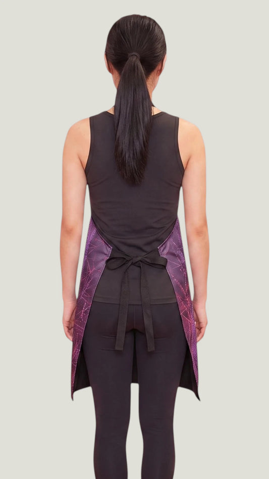 Back view of an Asian woman with a dark ponytail modeling the Ladybird Line 141 Rose Sparkle Waterproof & Bleach Resistant Apron, tied neatly at the waist over a black tank top and leggings against a light gray background.