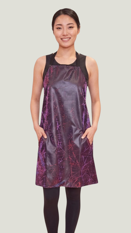 Front view of an Asian woman modeling the Ladybird Line 141 Rose Sparkle Waterproof & Bleach Resistant Apron, featuring a dark metallic finish with purple and pink sparkle line accents, deep side pockets, worn over a black top and leggings against a light gray background.