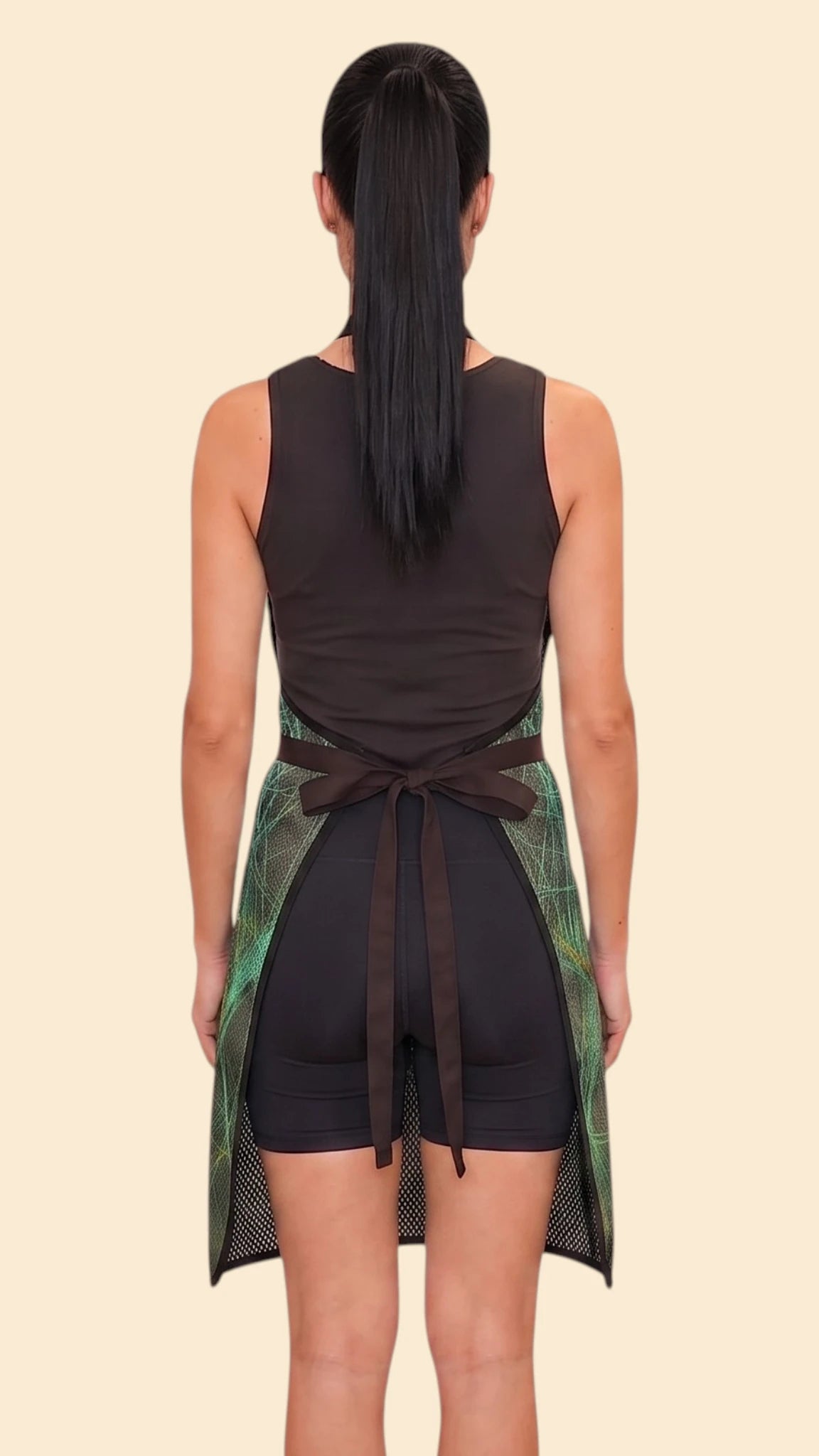 Back view of a woman with a dark ponytail modeling the Ladybird Line 142 Green Sparkle Waterproof & Bleach Resistant Apron, tied neatly at the waist over a black top and shorts against a pale beige background.
