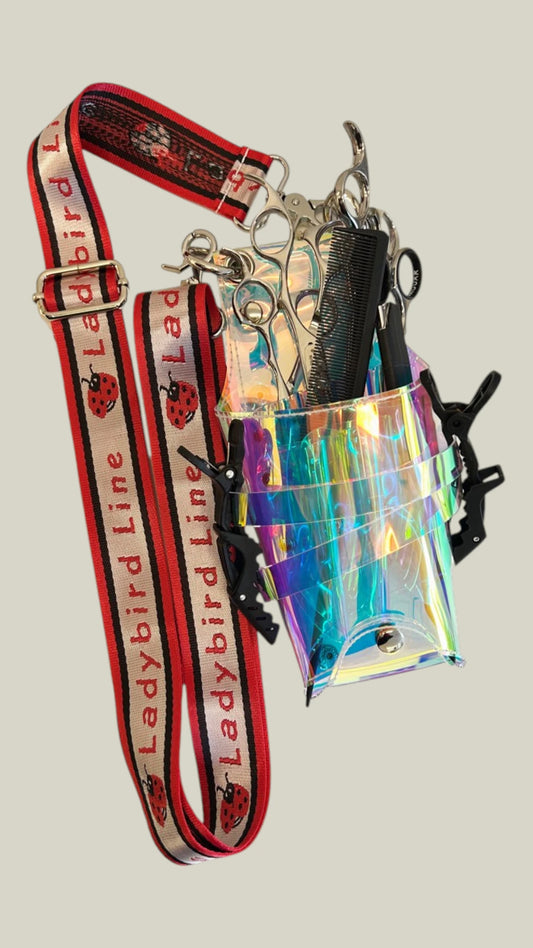 Iridescent clear holographic scissor caddy pouch filled with professional shears, combs, and clips, attached to a thick red adjustable shoulder strap printed with the Ladybird Line brand name and ladybug logos, set against a solid off-white background.