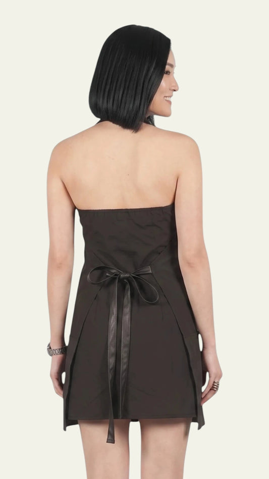 Back three-quarter view of a woman with short black hair modeling the Ladybird Line 150 Rhinestone Zipper Pro Water Resistant Salon Grooming Apron, showing the halter neck strap, open upper back design, and a black waist tie belt tied in a bow, set against a solid off-white background.