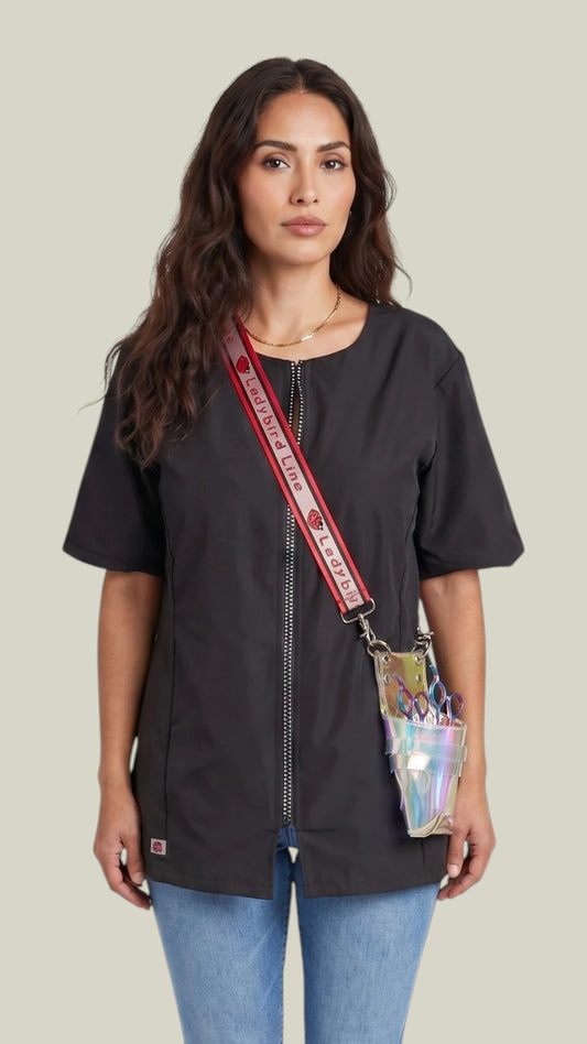 Front view of a woman with long dark wavy hair modeling the Ladybird Line 15 Crossover Body Scissor Case, featuring an iridescent clear scissor pouch worn cross-body with a red Ladybird Line branded strap, paired over a black short-sleeve jacket tunic and blue jeans against a solid light grey background.