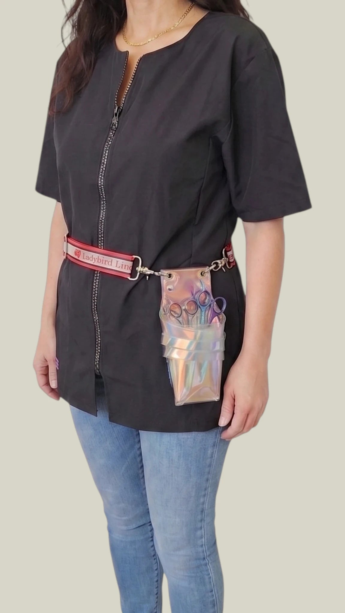Front view of a woman modeling the Ladybird Line 15 Crossover Body Scissor Case worn around the waist as a belt pouch, featuring a clear iridescent holographic scissor caddy filled with shears attached to a red Ladybird Line branded strap, paired over a black short-sleeve jacket tunic and blue jeans against a solid light grey background.