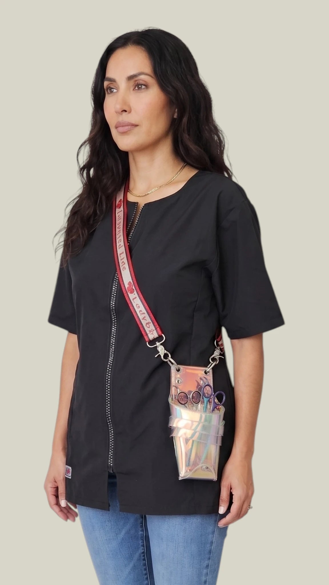 Side-view of a woman with long dark wavy hair modeling the Ladybird Line 15 Crossover Body Scissor Case, showing the clear iridescent holographic caddy pouch holding shears, worn cross-body over a black short-sleeve jacket tunic and blue jeans against a solid light grey background.